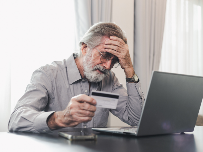 Older man clutches his forehead in anguish as he holds his credit card while sitting in front of his laptop and mobile phone