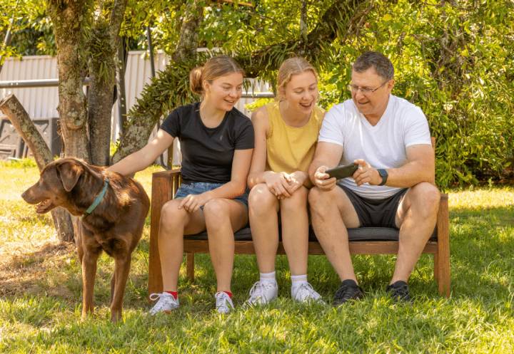 Family sitting on bench seat in their garden looking at a phone smiling with the pet dog nearby